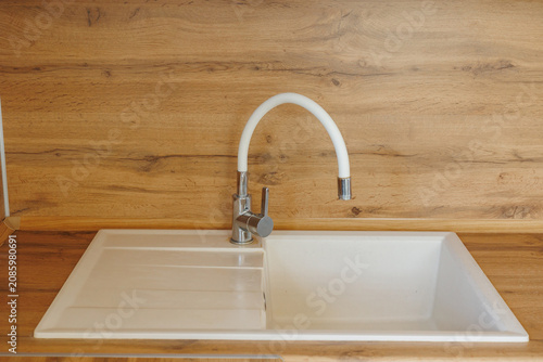 Curved white faucet rises over composite sink and built in drainboard beside chrome base and oak wood backsplash, creating clean modern kitchen utility with soft natural light and closeup detail