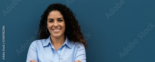 Smiling Hispanic woman with curly hair stands arms crossed in front of blue wall. Wears light blue shirt  looking confident  professional. Portrait conveys success  approachable demeanor for various
