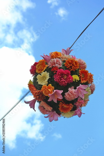 Farbenfrohe, runde Blumendekoration an einem Seil über einer Straße in der Altstadt von Quedlinburg, aufgenommen von unten vor einem strahlend blauen Sommerhimmel mit weißen Wolken.