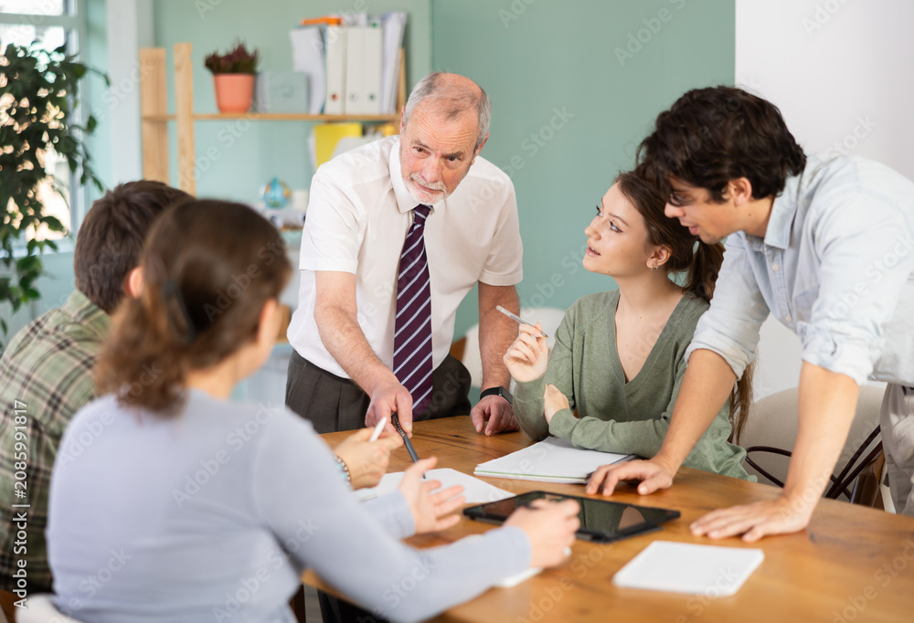 Fototapeta premium Elderly male professor talks to group of young students sitting at table and discussing assignment