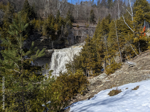 A scenic landscape view of Cataract Falls in Forks of the Credit Provincial Park. Heavy water flows over the rocky cliff surrounded by evergreen trees and melting winter snow under a clear blue sky.