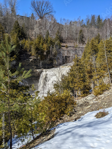 A scenic landscape view of Cataract Falls in Forks of the Credit Provincial Park. Heavy water flows over the rocky cliff surrounded by evergreen trees and melting winter snow under a clear blue sky.