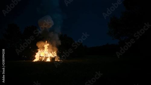 Small house burns in dark field at night. Flames and smoke create dramatic rural fire scene.