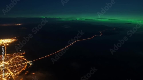 Illuminated modern city stretches across dark water under a vibrant green aurora