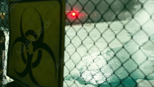 Warning sign, red emergency beacon, and contaminated liquid behind security fence create tense industrial biohazard scene, suggesting accident, spill, and hazardous release inside military laboratory.