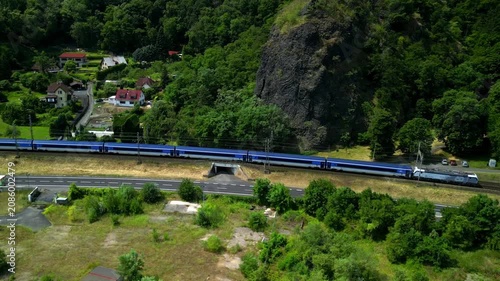Train travels along curved railway track beside lush green hillside and residential area, showcasing scenic landscape with trees and rock formations in the background