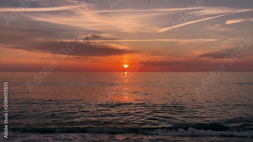 A tranquil, high-definition time-lapse capturing a vibrant, deep orange sunset over a rhythmic ocean. Gentle waves break against the shoreline, reflecting the brilliant colors of the setting sun, whil