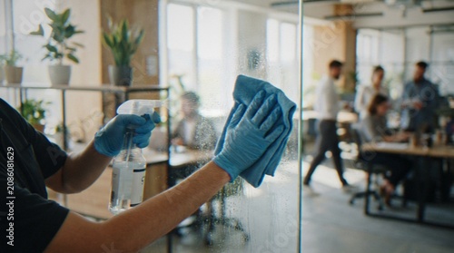 Bild auf Leinwand A janitor wearing blue nitrile gloves cleaning a glass office partition with a spray bottle and a cloth, with colleagues working in the blurred background
