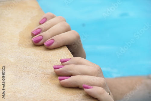 Fotografía Close-up of a woman's hands with pink painted fingernails holding onto the edge of a swimming pool
