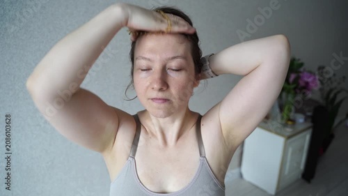 Static Shot Of Woman Adjusting Hair And Stretching Arms In Home Bedroom Interior During Morning Routine Beside Neutral Wall And Plant.