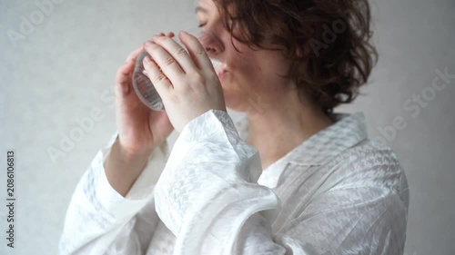 Static Shot Of Woman Drinking Water From Glass In White Robe During Morning Routine At Home Against Neutral Wall Background.