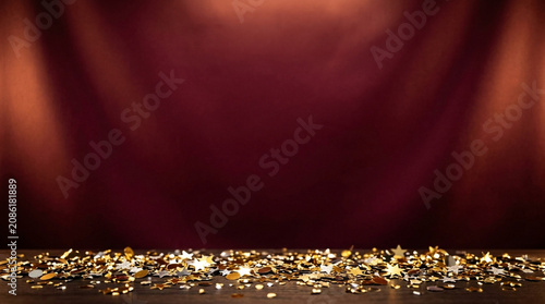 Golden and silver star-shaped confetti on a dark wooden surface against a maroon textured backdrop with soft overhead lighting, festive celebratory background for awards or events.