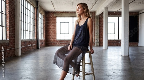 Thoughtful young woman sitting on rustic wooden stool inside spacious industrial brick loft