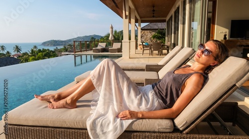 Relaxed young woman resting on sun lounger next to luxury infinity pool overlooking sea