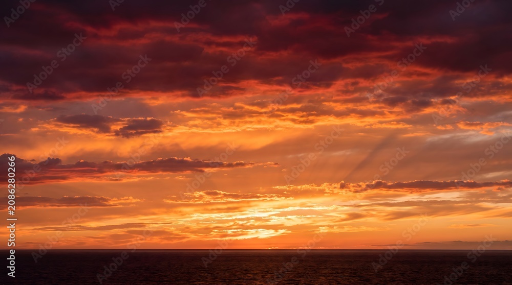 Fototapeta premium Dramatic fiery red and golden sunset sky over a dark ocean horizon. Breathtaking majestic clouds with glowing sun rays. Perfect nature landscape background for travel and weather concepts