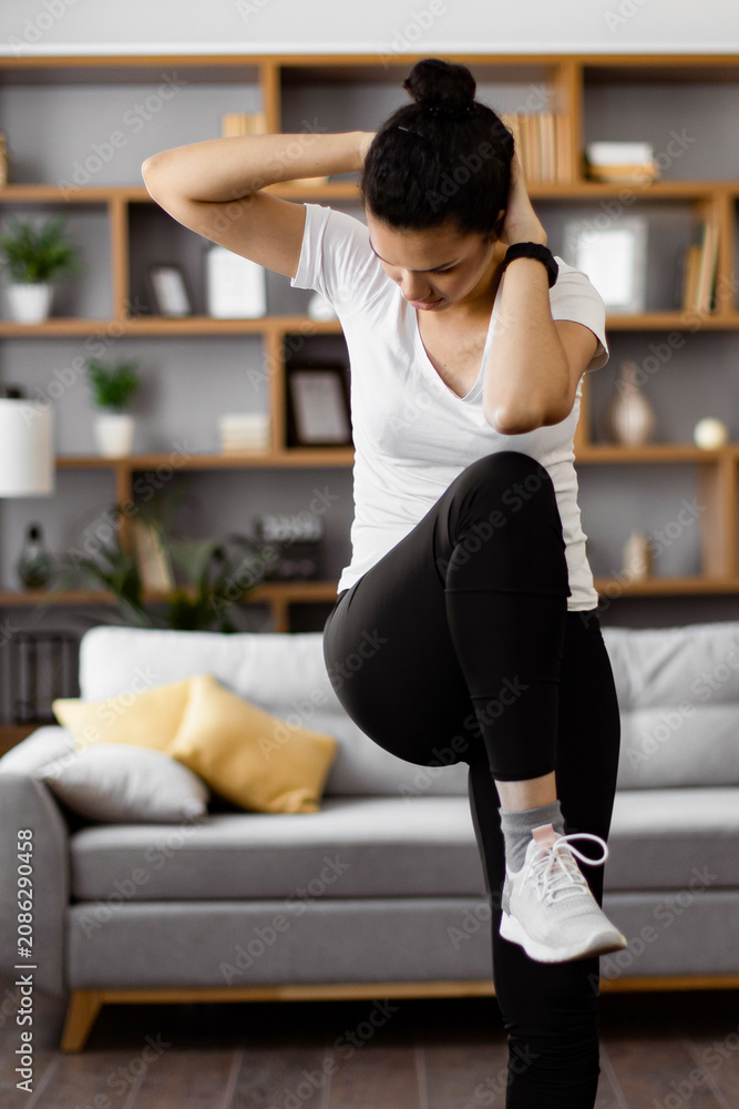 Obraz premium A young woman performs an abdominal crunch exercise while standing in her living room, focusing on her fitness routine