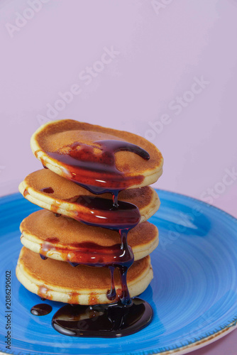 Freshly cooked pancakes stacked on a blue ceramic plate, dripping with sweet syrup against a light purple background