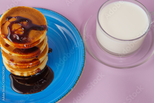 Stack of fluffy pancakes on a blue plate, drizzled with syrup, next to a glass of milk on a light purple surface