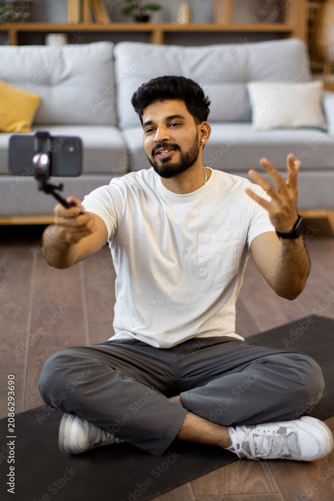Obraz premium A young Indian man sits cross-legged on a yoga mat, holding a smartphone on a selfie stick to record a video