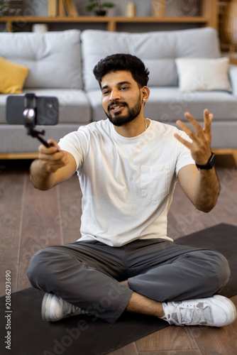 A young Indian man sits cross-legged on a yoga mat, holding a smartphone on a selfie stick to record a video