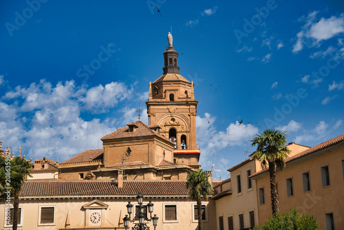 Ancient church tower rises above terracotta rooftops. Palm trees sway beside historic buildings. Bright blue sky holds scattered white clouds. Clock face glows softly on adjacent wall