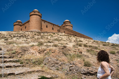 Woman in white walks toward ancient stone fortress. Bright blue sky frames rugged, sunlit hillside. Her curly hair catches golden afternoon light. Castle towers stand solemn against clear horizon