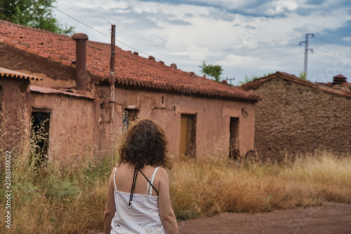 Woman stands before abandoned adobe buildings. Dried grass sways in the breeze. Overcast sky adds solemn mood. She gazes at empty, sun-baked structures. Her posture suggests quiet contemplation