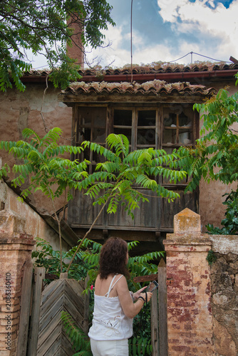 Woman stands before rustic wooden balcony. Green leaves frame weathered stone and brick walls. Her hand grips a gate, gaze fixed toward old window. Clouds drift above traditional tiled roof
