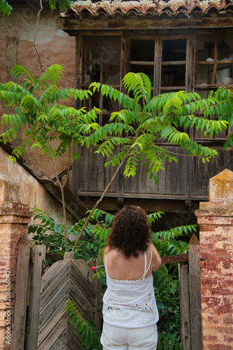 Woman stands before rustic wooden door, back to camera. Green leaves frame her, casting soft shadows. Weathered brick walls and tiled roof add character. Her calm posture suggests quiet contemplation