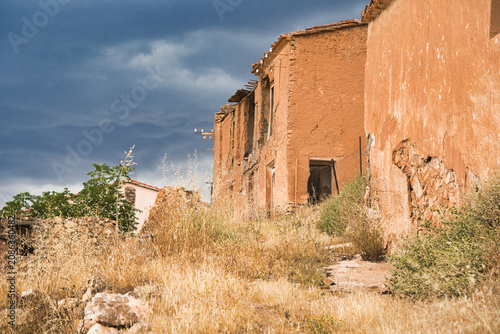 Abandoned adobe structure stands under a dramatic sky. Dry grasses and shrubs grow wild around crumbling walls. Weathered stone steps lead toward an empty doorway