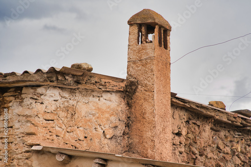 An old stone chimney rises above a weather-worn roof. Its rough texture evokes history and tradition. A cloudy sky frames the serene village scene. Cultural heritage captured in a simple, tranquil ima