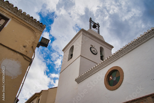 White church tower stands tall against blue sky. Clock face and bell mark sacred structure's presence. Clouds drift gently above ancient stone walls. Warm tones contrast cool sky