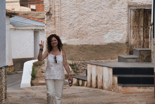Woman waves cheerfully in rustic village street. Warm sunlight bathes stone walls and narrow walkway. Her smile radiates friendliness and ease. Perfect for travel, lifestyle