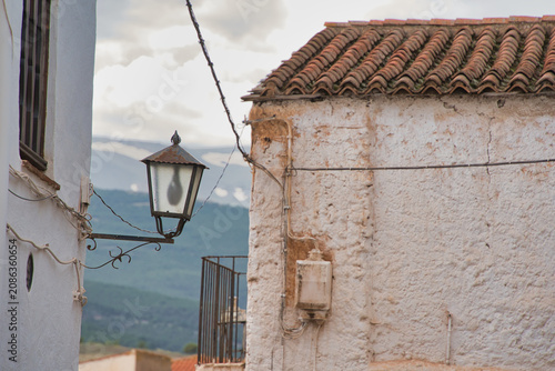 Weathered walls frame a rustic lamp. Red tile roof stretches across the scene. Mountains fade softly in the distance. Cable lines hint at old-world charm. Soft light glows over aged stone