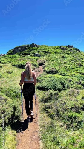 Fit woman hiking up a green mountain trail with trekking poles under blue sky