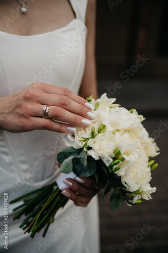 A slim, young and healthy bride in a white fitted wedding dress holds a white bouquet of roses with a gold wedding ring on her finger