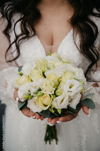 A slender, young, healthy bride in a white dress embroidered with pearls and a deep neckline with a large bust, holds a bouquet of white roses in her hands in front of her