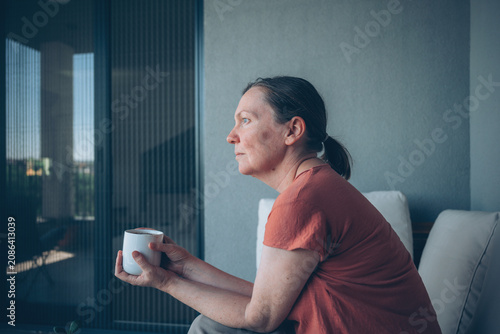 Fotografia Middle-aged woman sits on a balcony holding a coffee cup and gazes thoughtfully into the distance