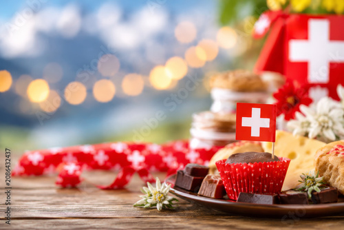 Swiss National Day celebration table with chocolate, cheese, and edelweiss flowers on alpine background.