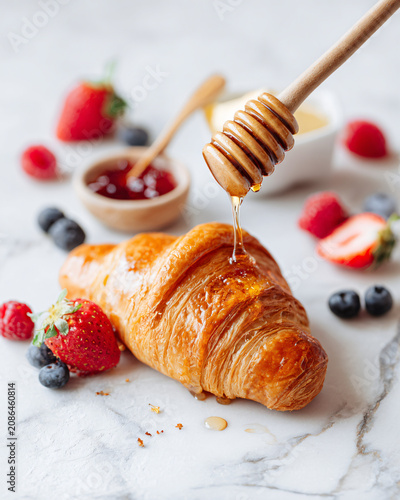 Golden butter croissant served with creamy European butter, artisan strawberry jam and organic honey dripping from a wooden honey dipper, fresh berries scattered naturally