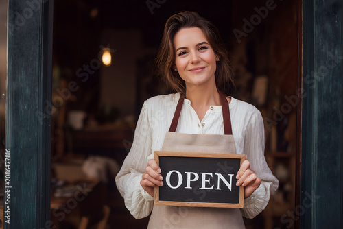 Woman waitress apron, cafe owner standing in the doorway, holding a sign with Open waiting for customers. Small business concept, cafes and restaurants