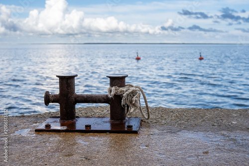 Photography Rusty mooring bollard with a weathered rope on a concrete pier overlooking a calm sea under dramatic summer clouds