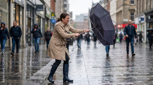 Woman battling wind and rain on city street, umbrella turning inside out, facing adverse weather conditions
