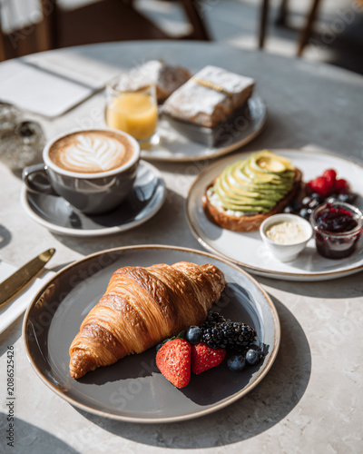 Luxury boutique café breakfast featuring buttery French croissant, avocado toast, cappuccino with latte art, artisan strawberry jam, creamy butter, fresh berries,