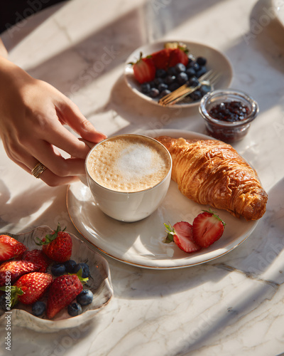 Elegant female hands gently placing a cappuccino beside a warm flaky butter croissant, creamy butter, artisan berry jam, fresh strawberries and blueberries, white marble breakfast table