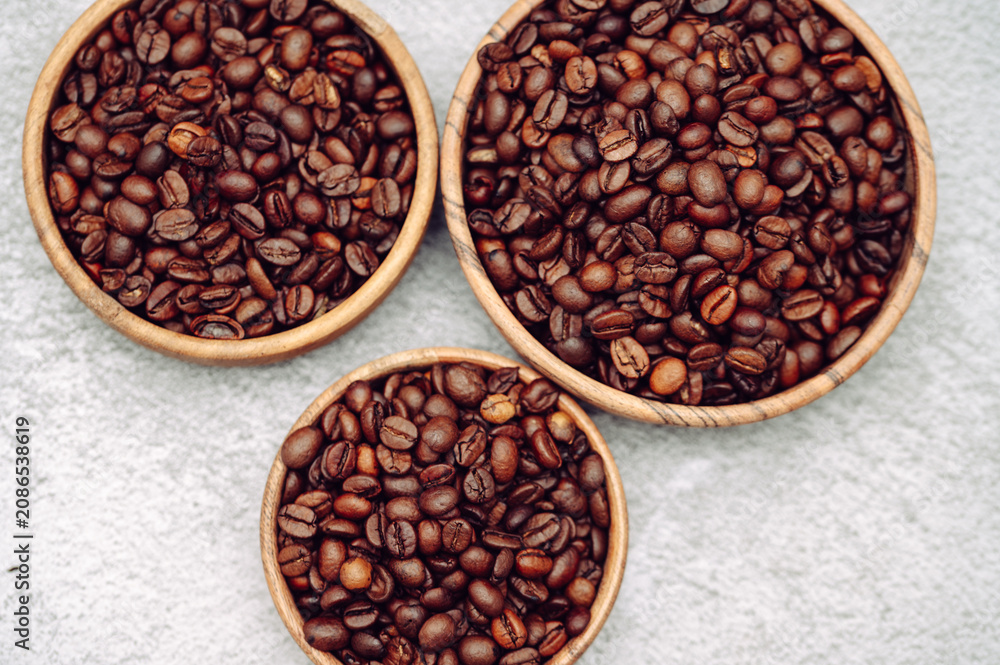 Fototapeta premium Three round wooden bowls filled with roasted brown coffee beans on a grey concrete background.Aromatic dark roast coffee beans arranged in circular wood plates, top view copy space.
