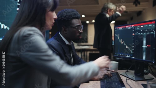 African American male broker typing on keyboard while watching falling financial chart. Female colleague sitting beside him in dark trading office. Senior manager standing behind team at night.