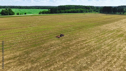 Tractor harvesting grass in a field marked with cut rows under a bright blue sky, aerial view