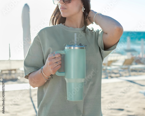 Blank sage green 40oz tumbler mockup, young woman at beach at sunset, hands closeup. Lifestyle scene