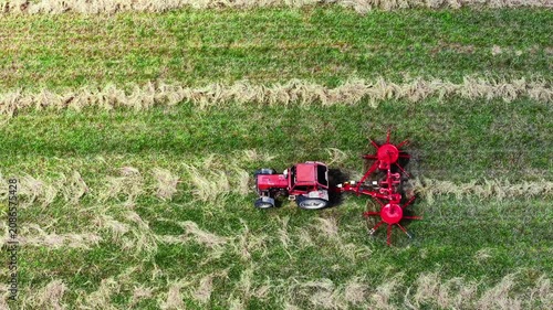 Aerial view of Tractor with red rake working on green pasture field creating neat furrows during harvest season